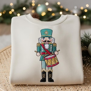 May include: Cream-colored sweatshirt featuring an embroidered nutcracker soldier. The nutcracker is depicted in teal, red, gold, and black, holding drumsticks and a drum. The design is centered on the front of the sweatshirt.