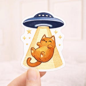 May include: Embroidered patch featuring a cartoon orange cat being abducted by a blue UFO. The cat is smiling and floating in a beam of light. Gold stars surround the UFO and cat.