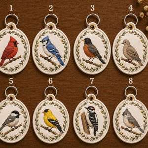 May include: Eight embroidered bird keychains. Each oval keychain features a different bird design, including a cardinal, blue jay, robin, dove, chickadee, goldfinch, woodpecker, and tufted titmouse. The birds are perched on branches within a floral wreath.