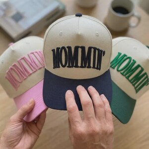 May include: Three baseball caps with the word "MOMMIN'" embroidered on the front. The caps have a cream-colored crown and a navy blue brim. One cap has pink embroidery and brim, another has green embroidery and brim, and the third has navy embroidery and brim.