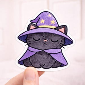 May include: Embroidered patch of a black cat in a purple wizard hat with yellow stars and a matching cape. The cat has closed eyes and a pink nose. The patch has a white border, creating a whimsical design.