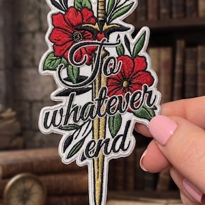 May include: Embroidered patch featuring a gold sword, red flowers, and green leaves. The patch has the text "To whatever end" in a stylized font. The patch is held by a person with pink nail polish.
