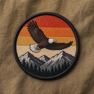 May include: Embroidered patch depicting a bald eagle in flight against a sunset. The design includes a mountain range and forest at the bottom, with a black border. The eagle is brown and white, with a yellow beak and talons.