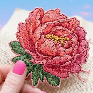 May include: A detailed, hand-stitched peony flower embroidery patch. The flower features layers of pink and coral thread with green leaves and yellow stamens. The patch is held against a neutral fabric background.