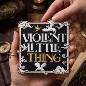 May include: Black square patch with the words "VIOLENT LITTLE THING" in white and gold lettering. The patch features dragon illustrations and a gold and white border. The patch is held in a person's hands.