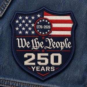 May include: A navy blue and red embroidered patch in the shape of a shield. The patch features the American flag, the text "We the People", and the numbers "250 YEARS" with the years "1776-2026" in a circle.