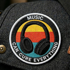 May include: A circular embroidered patch featuring a black pair of headphones centered over a horizontal striped background of yellow, orange, and teal. The black border contains the white text MUSIC CAN CURE EVERYTHING in a bold sans-serif font.