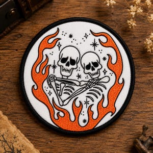 Peut inclure: Un écusson brodé circulaire représentant deux squelettes blancs enlacés, entourés de flammes orange et de petites étoiles noires sur un fond blanc avec une bordure noire épaisse. Le motif est centré et symétrique.