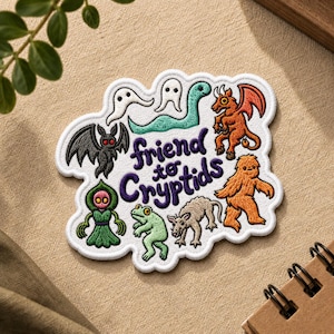 May include: Embroidered patch featuring various cryptids, including a bat, ghost, dragon, and Bigfoot. The patch has the text "friend to cryptids" in purple. The patch is white with a stitched border and is on a tan surface.