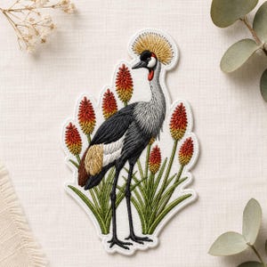 May include: Embroidered patch featuring a Grey Crowned Crane with a black head, red wattle, and yellow crown. The crane stands among red and yellow flowers and green grass. The patch has a white border.