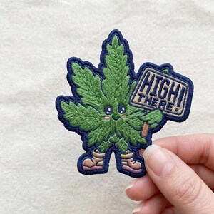 May include: Embroidered patch featuring a smiling green cannabis leaf character with a blue outline. The character holds a sign that reads "HIGH THERE!" in a rectangular frame. The patch has a cute, cartoonish design with pink blush.