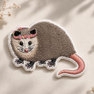 May include: An embroidered patch featuring a gray opossum with a white face, black ears, and a pink nose. The opossum wears a colorful floral crown made of small pink, yellow, and orange flowers. It has a long, curved pink tail and black feet.