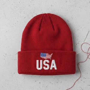 May include: A red knit beanie with a white embroidered "USA" text and a small American flag graphic. The hat is made of a soft, ribbed material, perfect for cold weather. The beanie is a classic style.