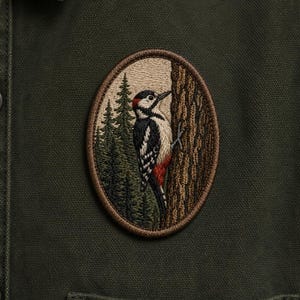 May include: An embroidered patch featuring a woodpecker clinging to a tree trunk, set against a backdrop of pine trees. The patch has a brown border and is sewn onto a dark green fabric, likely a shirt or jacket.