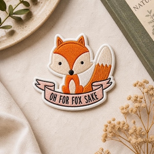 May include: Embroidered patch featuring a cartoon fox in orange, white, and tan. The fox sits above a pink banner with the text "OH FOR FOX SAKE" in black. The patch has a white border.