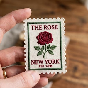 May include: Embroidered patch in the shape of a postage stamp. The patch features a burgundy rose with green leaves, the text "THE ROSE" at the top, and "NEW YORK" and "EST. 1788" at the bottom, all in burgundy.