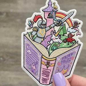 May include: Embroidered patch featuring a knight, unicorn, and other fairytale characters emerging from an open book. The book is purple with the words "Fairy Tales" on the spine. A rainbow arches over a castle.