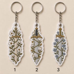 May include: Three embroidered keychains featuring silver-toned metal rings and chains. Each keychain displays a sword entwined with floral vines. The first has white flowers, the second has white and yellow flowers, and the third has blue flowers.
