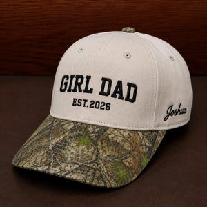 May include: A beige baseball cap with a camouflage patterned brim. The front features black embroidered text reading GIRL DAD EST. 2026. The side of the cap displays the name Joshua in black script embroidery. The top button is also camouflage.