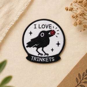 May include: Embroidered patch featuring a black crow holding a red gem in its beak, with the text "I LOVE TRINKETS." The crow is set against a white background with black accents and a black border. The patch is likely for clothing or accessories.
