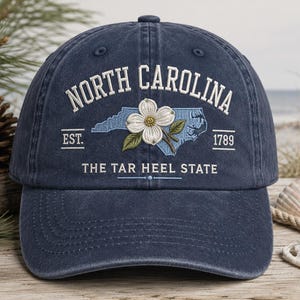 May include: A navy blue baseball cap with embroidered text and a floral design. The cap features the words "NORTH CAROLINA" above a light blue outline of the state, a white flower, and the text "THE TAR HEEL STATE". The year "1789" is also displayed.