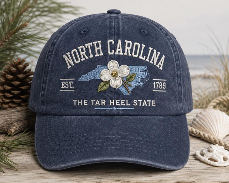 May include: A navy blue baseball cap with embroidered text and a floral design. The cap features the words "NORTH CAROLINA" above a light blue outline of the state, a white flower, and the text "THE TAR HEEL STATE". The year "1789" is also displayed.