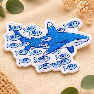 May include: Embroidered patch depicting a blue shark with smaller blue and white fish. The patch has a white border and is made with detailed stitching, giving it a textured look. The design is set against a neutral background.