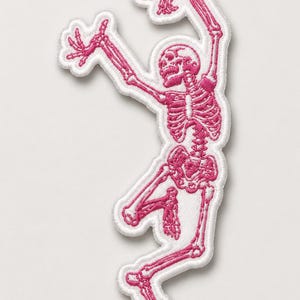 May include: Embroidered patch depicting a pink skeleton in a dancing pose. The skeleton is detailed with visible ribs, skull, and limbs, with a white border. The patch is designed for application on clothing or accessories.