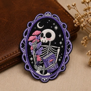 May include: Embroidered patch featuring a skeleton holding flowers and a book within a purple oval frame. The skeleton has a smiling skull and is set against a black background with stars and a crescent moon. The frame has a scalloped edge.