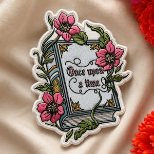 May include: Embroidered patch featuring a storybook design with the words "Once upon a time." The book is light blue with gold accents, surrounded by pink flowers and green vines. The patch has a white border.