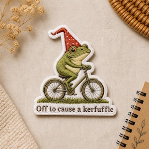 May include: Embroidered patch featuring a green frog riding a bicycle and wearing a red polka-dot hat. The patch has the text "Off to cause a kerfuffle" in brown lettering. The patch is white with a green and brown design.