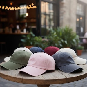 May include: A collection of baseball caps in various colors, including pink, green, navy, and burgundy, are displayed on a round wooden table. The caps have a classic design with a curved brim and adjustable strap. The background is blurred, suggesting an outdoor setting.