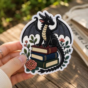 May include: An embroidered patch featuring a black dragon with cream-colored underbelly scales perched atop a stack of three books in red, blue, and green. The design is framed by green leafy stems, small pink flowers, and a large peach-colored rose.