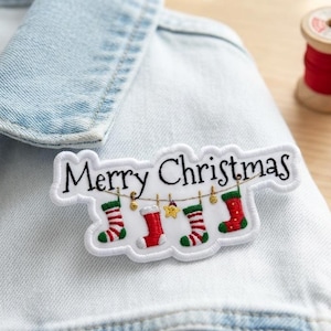 May include: A white embroidered patch featuring the black text Merry Christmas above a string of four colorful Christmas stockings and a small yellow star. The patch is attached to a light blue denim fabric with visible stitching details.