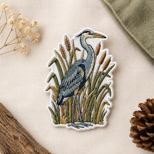 May include: Embroidered patch featuring a blue heron standing among cattails and tall grass. The bird has a long neck and beak, with detailed feathering. The patch is bordered with white stitching, and is a nature-themed item.