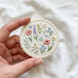 May include: A round, embroidered patch featuring a floral design. The patch showcases a variety of flowers in shades of pink, blue, lavender, and cream, with green leaves. The patch has a scalloped edge and is set against a white background.