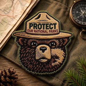 May include: Embroidered patch of a bear wearing a ranger hat, with the text "PROTECT OUR NATIONAL PARKS." The patch is brown, tan, and green, with detailed stitching. A map, compass, and pine needles are in the background.