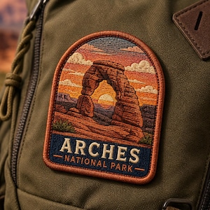May include: Embroidered patch depicting a sunset over a rock formation in Arches National Park. The patch is rectangular with a brown border and the words "ARCHES NATIONAL PARK" in white text on a navy blue background. It's attached to a green backpack.