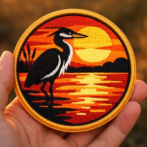 May include: Embroidered patch featuring a black heron standing in water with a sunset backdrop. The heron has white accents and a long beak. The sunset is a gradient of orange and yellow, with a black silhouette of land. The patch has a gold border.