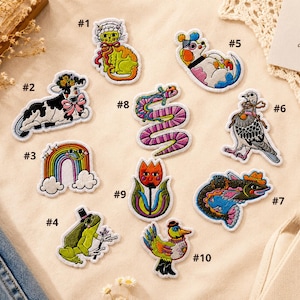 May include: A collection of ten embroidered patches featuring various designs: a cat, a cow, a rainbow, a frog, a dog, a pigeon, a fish, a snake, a tulip, and a bird. Each patch is numbered from one to ten and displays colorful, detailed stitching.