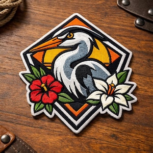 May include: Embroidered patch with a heron design. The patch features a heron with a long orange beak, grey and white feathers, and a yellow background. It includes red and white flowers with green leaves, set within a black and white diamond shape.