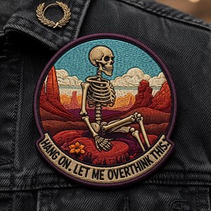 May include: Embroidered patch featuring a skeleton in a desert landscape with the text "HANG ON. LET ME OVERTHINK THIS." The patch is circular with a purple border and is sewn onto a black denim jacket. A gold-colored laurel wreath pin is also visible.