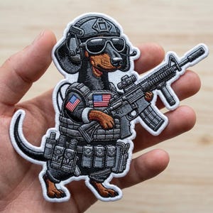 May include: Embroidered patch featuring a dachshund dog in military gear, including a helmet, sunglasses, and tactical vest with American flag patches. The dog is holding a rifle. The patch has a white border.