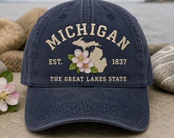 Op de afbeelding: Een marineblauwe baseballcap met geborduurde tekst en afbeeldingen. De tekst luidt "MICHIGAN" boven een kaartomtrek en bloemen, met "EST. 1837" en "THE GREAT LAKES STATE" eronder. De cap is een klassieke stijl.