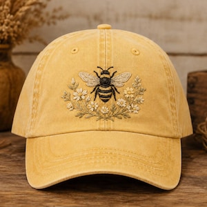 May include: A mustard yellow baseball cap featuring an embroidered design on the front panel. The embroidery depicts a detailed black and yellow bee centred above a semi-circular arrangement of small white and yellow flowers with green stems.