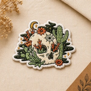 May include: Embroidered patch featuring a skull with desert flora. The skull is cream-colored, surrounded by green cacti, orange flowers, and a crescent moon. A spiderweb is embroidered on the skull. The patch has a white border.