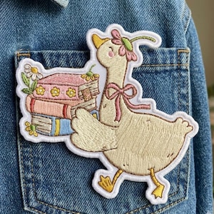 May include: Embroidered patch featuring a whimsical goose carrying a stack of books. The goose is cream-colored with a pink bow and a flower crown. The books are pink, blue, and yellow, with floral accents. The patch is on a denim jacket pocket.