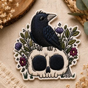 May include: Embroidered patch featuring a black crow perched atop a skull, surrounded by purple and maroon flowers and green leaves. A small spider web is visible on the lower right. The patch has a cream-colored background.