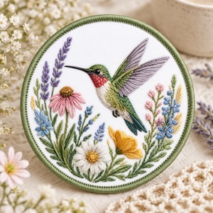 May include: Embroidered round patch featuring a hummingbird in flight, surrounded by colorful flowers and foliage. The hummingbird has green, red, and white feathers. The patch is bordered by a green stitched edge.