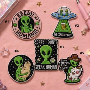 May include: A collection of embroidered patches with alien-themed designs. One patch features an alien with the text "EEEW! HUMANS!". Other patches include aliens in a UFO, with a cat, and with a cow. The patches are in various colors.